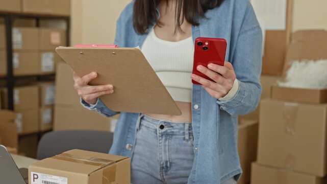 Woman holds clipboard and red smartphone, bare midriff visible, counts parcels and scans labels in a building; small business busy.