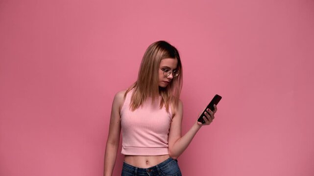 Annoyed young woman holding a smartphone and reacting to social media content on a pink background