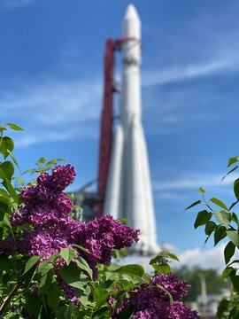Lilac blooms and bright green leaves in sharp focus with the vostok rocket monument softly blurred behind, blending moscows space heritage with springtime color