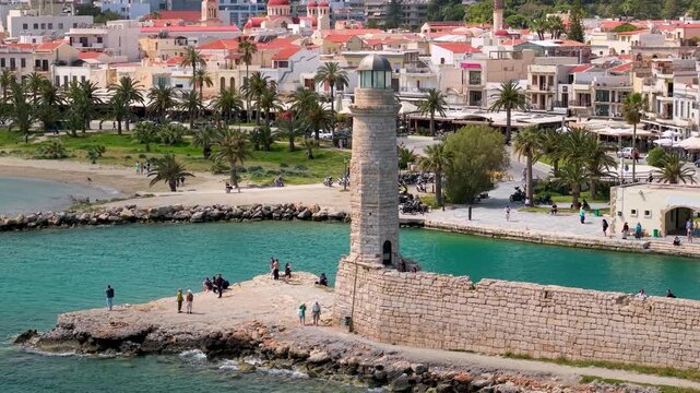 Rethymno Crete Greece 4k aerial drone pull back from Venetian lighthouse. Old town skyline with pink domes church and mosque minaret in background. Historical architecture of Rethymnon harbor