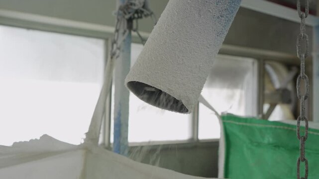 Industrial pipe outlet pouring recycled plastic pellets into green bulk bag suspended by chains