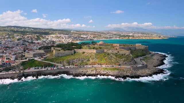 Aerial back flight from Rethymno fortress walls to old town and sea waves, Crete, Greece. Drone view of Venetian Fortezza, coastal road and historic city center at sunny day. Scenic landscape, 4k