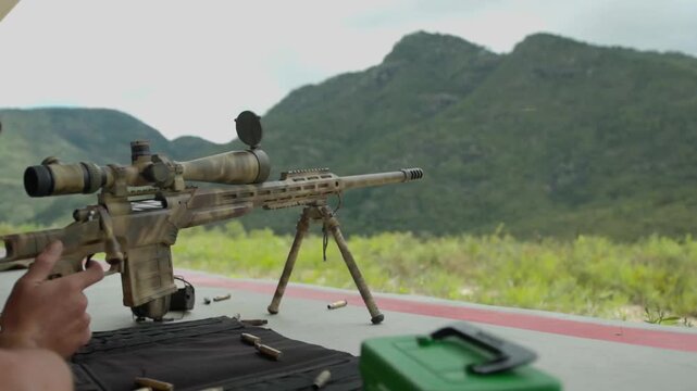 A sniper with his gun set up on top of a range with bullet shells on the ground around, waits for his shot and pulls the trigger, as the gun fire it lets out a puff of smoke.