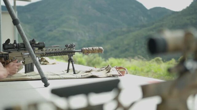 A sniper is laying down with gear set up around him, waits for his shot and fires his rifle as the bullet casing flies out and bounces in slow motion on the ground, with the mountains in background.