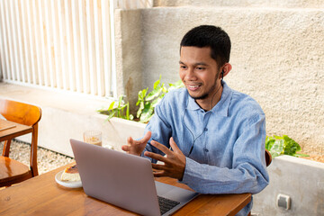 Man smiles while video calling using laptop at outdoor cafe. He wears blue shirt and earphones....