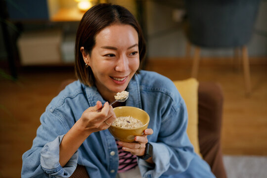 Happy Asian Korean woman eating oatmeal while relaxing at home
