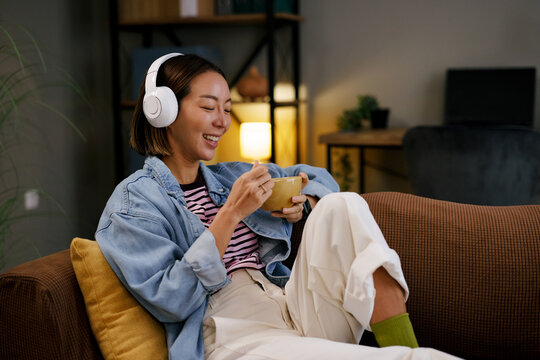 Smiling asian woman with headphones eating breakfast on sofa at home