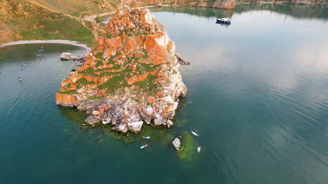 Lake Baikal on a summer evening. A flight over Cape Burkhan or Shamanka Rock on Olkhon Island. Tourists paddleboard in the bay. Active recreation.