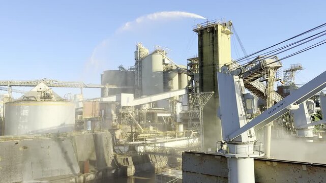 Aerial view of grain terminal with own berth. Bulk cargo ship docked while grain flows through loading arms. Huge clouds of grain dust and industrial water spray create dramatic port scene.
