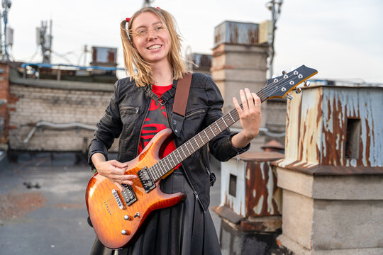 Young smiling woman with dreadlocks holding electric guitar on urban rooftop, cheerful rock musician outdoor lifestyle concept.