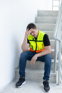 Exhausted and stressed male worker sitting heavily on concrete stair wearing yellow safety vest at local construction site expressing feelings of deep frustration and being completely tired