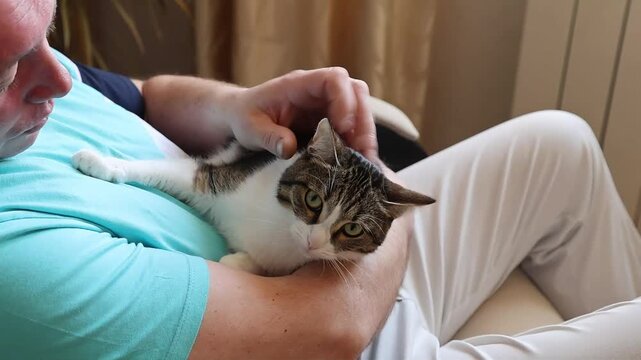 A middle-aged man relaxes in an armchair, tenderly holding a cat in his arms, exuding calm and affection