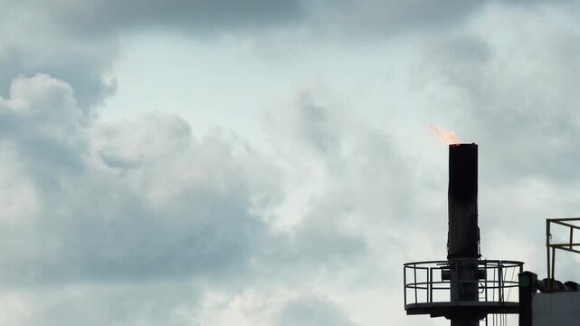 Industrial gas flare chimney with fire burning against a cloudy sky. Concept of energy production, pollution, and oil industry.