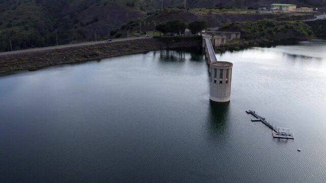 Scenic aerial view flying over a water reservoir intake tower and surrounding green hills reflecting on the surface