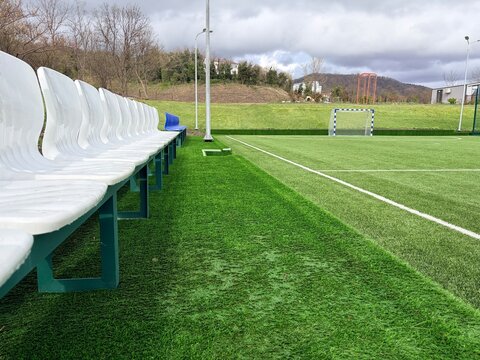 A soccer field, featuring a row of white plastic stadium seats with green metal frames running along the left edge.