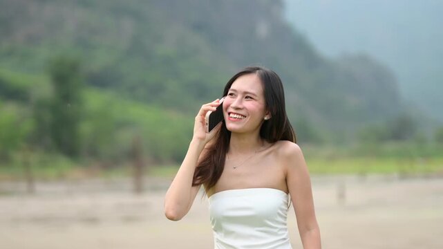 A young woman in a white strapless dress talks on her phone outdoors with a blurred natural background.