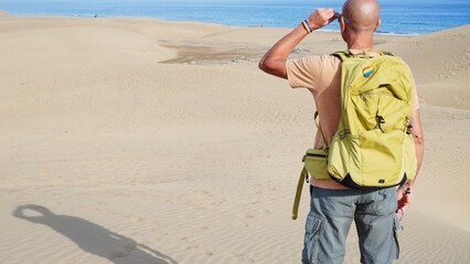 Man with a rainbow-flag backpack and pride bracelet stands on Maspalomas sand dunes, gazing at the Atlantic, embracing summer freedom and LGBTQ+ identity beside the sea © Canary4stock