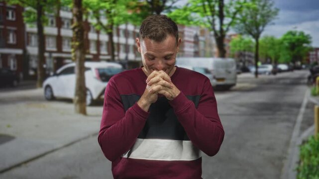 Man clasping hands with interlaced fingers and smiling on a residential urban street with parked cars and trees; amusement anticipation playfulness.