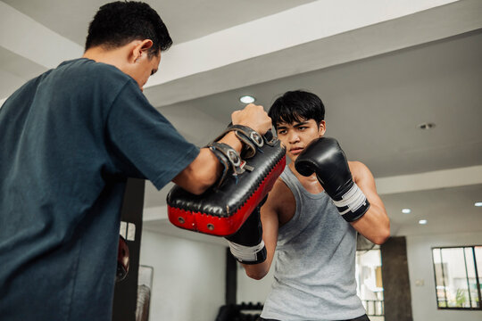 Focused young boxer delivers an uppercut to Thai pads held by coach during intense boxing training session indoors.
