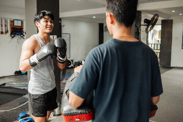 Naklejka premium Young Asian boxer playfully sparring with coach holding Muay Thai pads, preparing for striking drills in a gym.