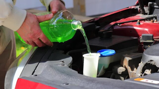 Close-up of hands pouring summer windshield washer fluid into reservoir under car hood, refilling screen wash liquid in engine bay outdoors, car maintenance and care, vehicle service routine, auto