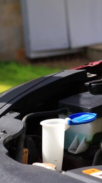 Close-up of hands pouring summer windshield washer fluid into reservoir under car hood, refilling green screen wash liquid in engine bay outdoors, car maintenance and care, vehicle service routine and