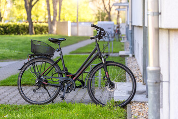 Fototapeta premium Black bicycle parked residential building area green lawn paved walkway quiet urban neighborhood Germany. Modern city bike basket rack stands sunlight creating calm everyday commuting atmosphere