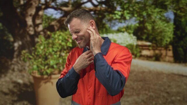 Man in orange safety uniform clutches his ear with one hand and presses his neck with the other, squinting in pain on street; earache pain.