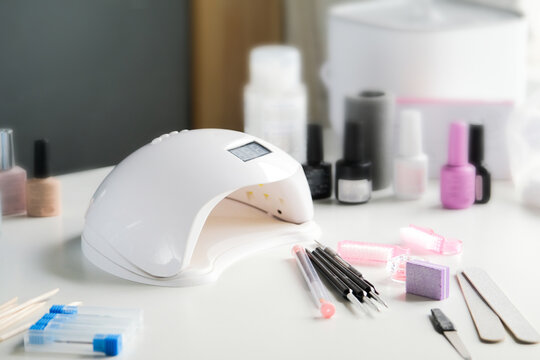 Nail LED lamp, colour gel polishes and other accessories on white table.
