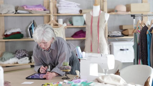 Mature dressmaker draws a sketch of clothes and patterns on an electronic tablet. Woman creates a fashion design in a workshop.