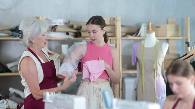 Skilled seamstress with a needle on arm sews fabric against the background of a mature atelier worker who shows a sample to a client. High quality 4k footage