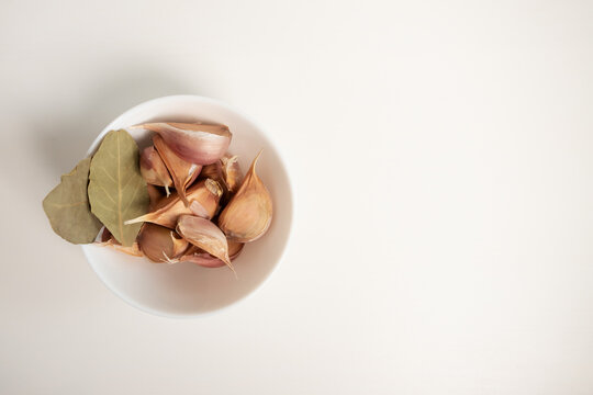 garlic cloves with bay leaves in a white bowl on a light background, styled as a minimal aromatic ingredient set for meat, rice and homemade cooking recipes