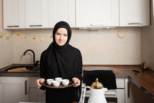 smiling muslim woman in black hijab holding a copper tray with white finjan cups for arabic coffee serving in a bright home kitchen