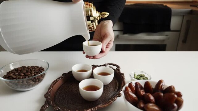 arabic coffee being poured from white dallah into a finjan cup on a copper serving tray with filled cups, dates and cardamom on the table