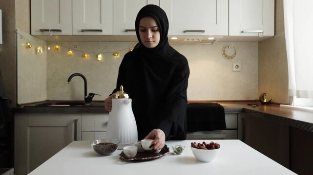 muslim woman in black hijab pouring arabic coffee from white dallah into a small finjan cup beside dates and cardamom on the table at home