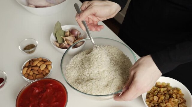 close-up of a spoon lifting long grain basmati rice from a large glass bowl surrounded by garlic bay leaves almonds raisins and tomato sauce for a richly seasoned rice recipe