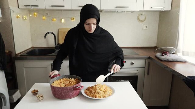 muslim woman in black hijab spoons fluffy cooked rice from a red pot onto a white serving plate, shaping the base for a generous homemade family style main course