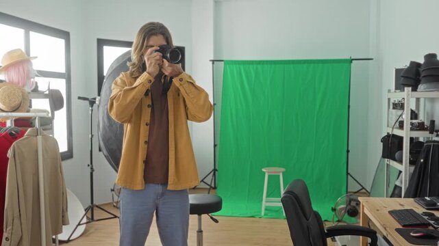 Man holds camera to eye with hands on lens and shutter, standing before a green screen and studio lights and stool visible; concentration creativity.