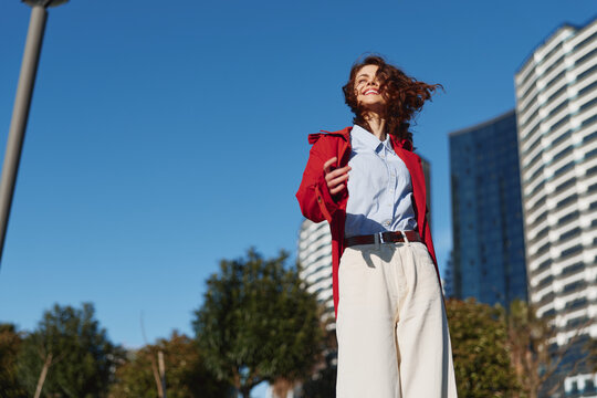 A young woman with curly hair stands in an urban outdoor setting, wearing a red jacket over a white shirt and beige pants, smiling confidently as the city skyline rises behind her.