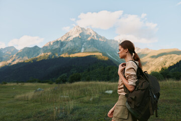 Fototapeta premium A young woman with a backpack stands in a grassy field looking at a mountain peak under a partly cloudy sky. Concept of outdoor adventure and lifestyle.