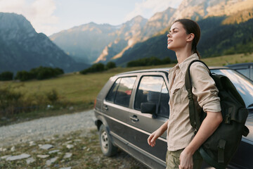 Fototapeta premium Young woman with a backpack walks beside a parked car in a rugged mountain valley, enjoying a bright crisp outdoor moment.