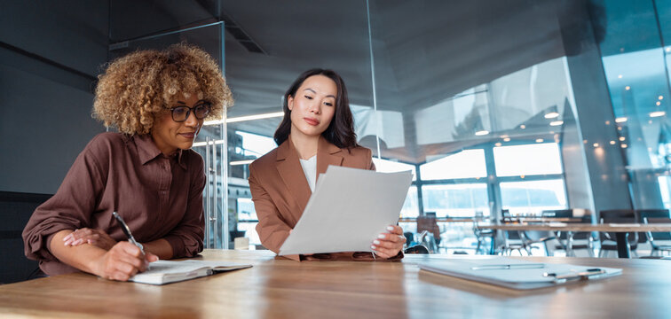 Two professional women businesswomen in modern office reviewing document, one taking notes at table, business meeting, teamwork, coaching and analysing paperwork collaboration. Banner, copy space