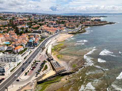 Aerial view of Avenida Marginal winding along the rocky coastline with waves crashing on the shore and urban buildings under a cloudy sky in Parede, Lisboa, Portugal.