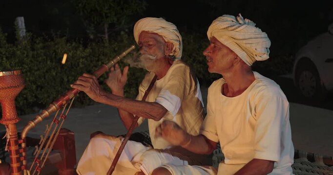 Indian old aged man wear white kurta pajama sitting on woven cot bed enjoy desi life at rural area. Happy two male group talk speak share gossip do hookah hold pipe spend night time village place