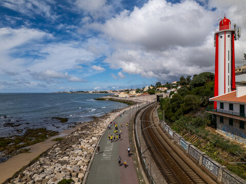 Aerial view of Passeio Maritimo de Alges with people exercising next to a red and white lighthouse and railway tracks along the coast in Cruz Quebrada, Lisboa, Portugal.