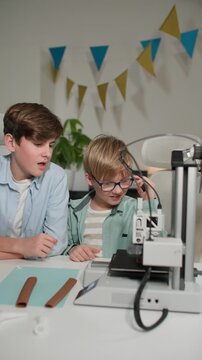 At a bright table, two schoolchildren watch a 3D printer work and play with it