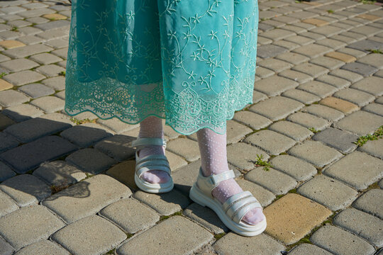 legs of a little girl in sandals in a dress, little girl standing on the cobblestones in a turquoise dress and white sandals, view below the knees