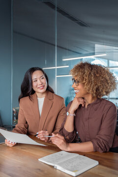 Two smiling businesswomen collaborate at desk in modern office, reviewing business financial tax document, conveying teamwork, mentoring and workplace communication. Multiracial team working. Vertical