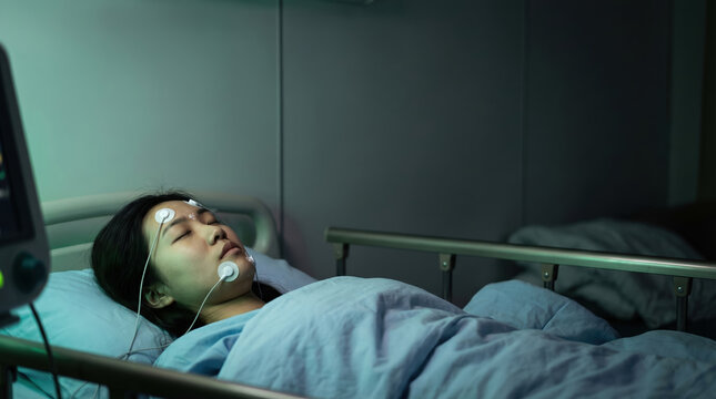 Asian woman undergoing sleep study test at clinic. Patient with sensors on face monitor breathing for snoring and apnea diagnosis. Healthcare sleep medicine and medical examination.