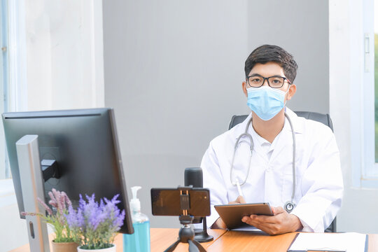 Portrait of a doctor sitting at a desk in an office with assorted digital devices wearing a face mask and laboratory coat